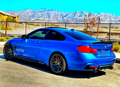 A blue sports car is parked on a sunlit road. It features dark wheels and a rear spoiler, with mountains and a clear sky in the background.