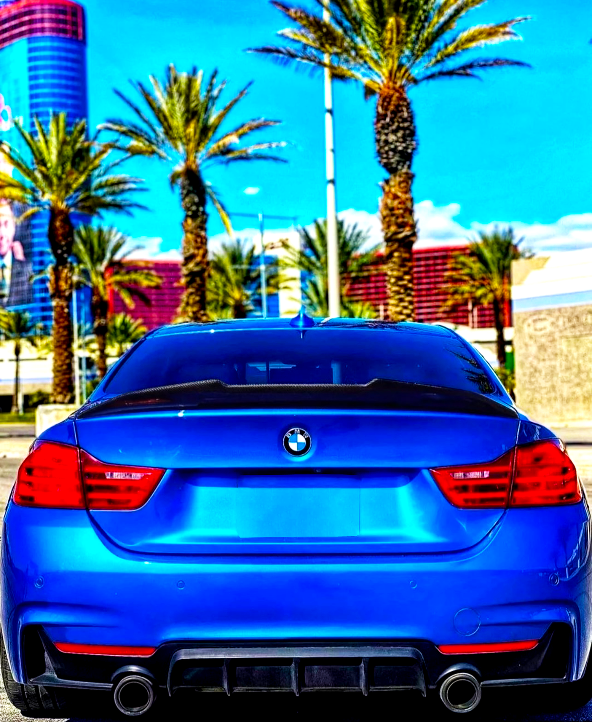 A blue BMW car is parked, facing a row of tall palm trees under a bright blue sky, with a modern building in the background.