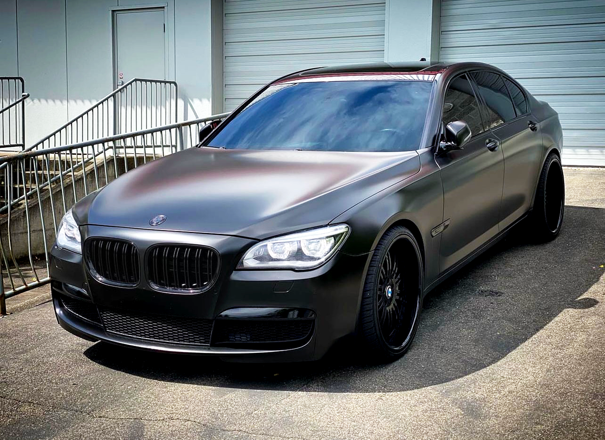 A sleek, matte black luxury sedan is parked on a paved surface. The vehicle is in front of a building with closed garage doors and a metal railing.