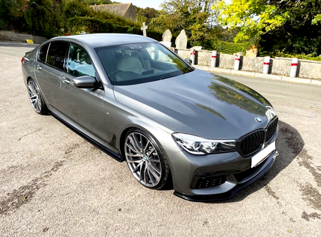A sleek, grey sedan is parked on a quiet road beside stone markers and lush greenery, under a clear sky.