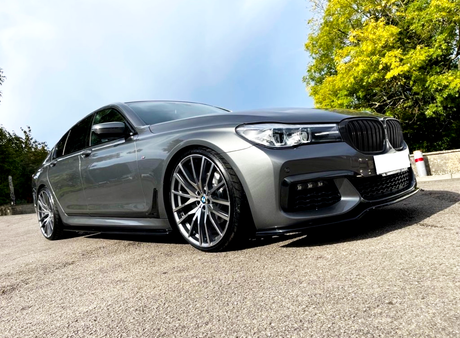 A sleek grey car parked on a paved surface with large alloy wheels, surrounded by greenery on a clear day.