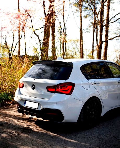 A white hatchback car is parked on a forested road, surrounded by tall trees and sunlight filtering through the branches. The rear lights are illuminated.