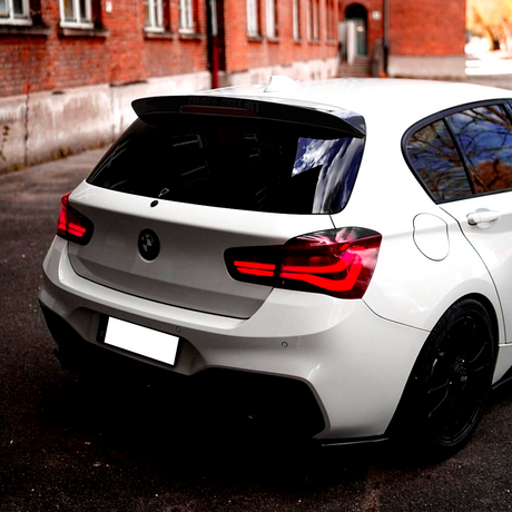 A white car with red tail lights is parked on a dark asphalt surface, surrounded by brick buildings reflecting in its windows. The rear license plate is blank.