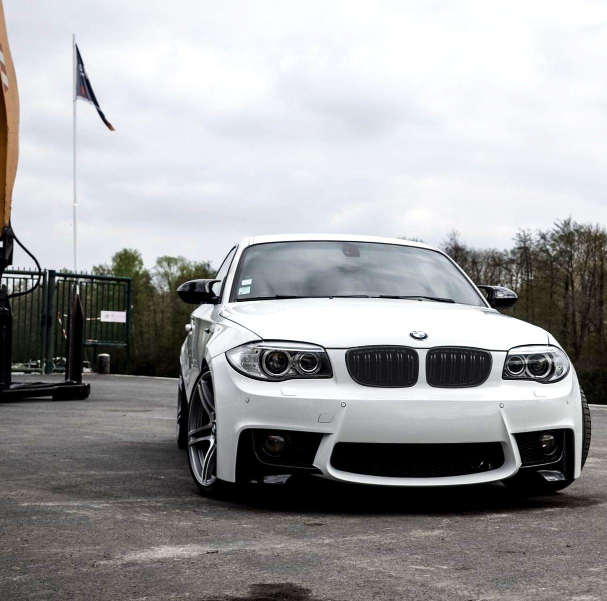 A white BMW car is parked on a paved surface near a green fence and trees. An American flag flies on a pole in the background under a cloudy sky.