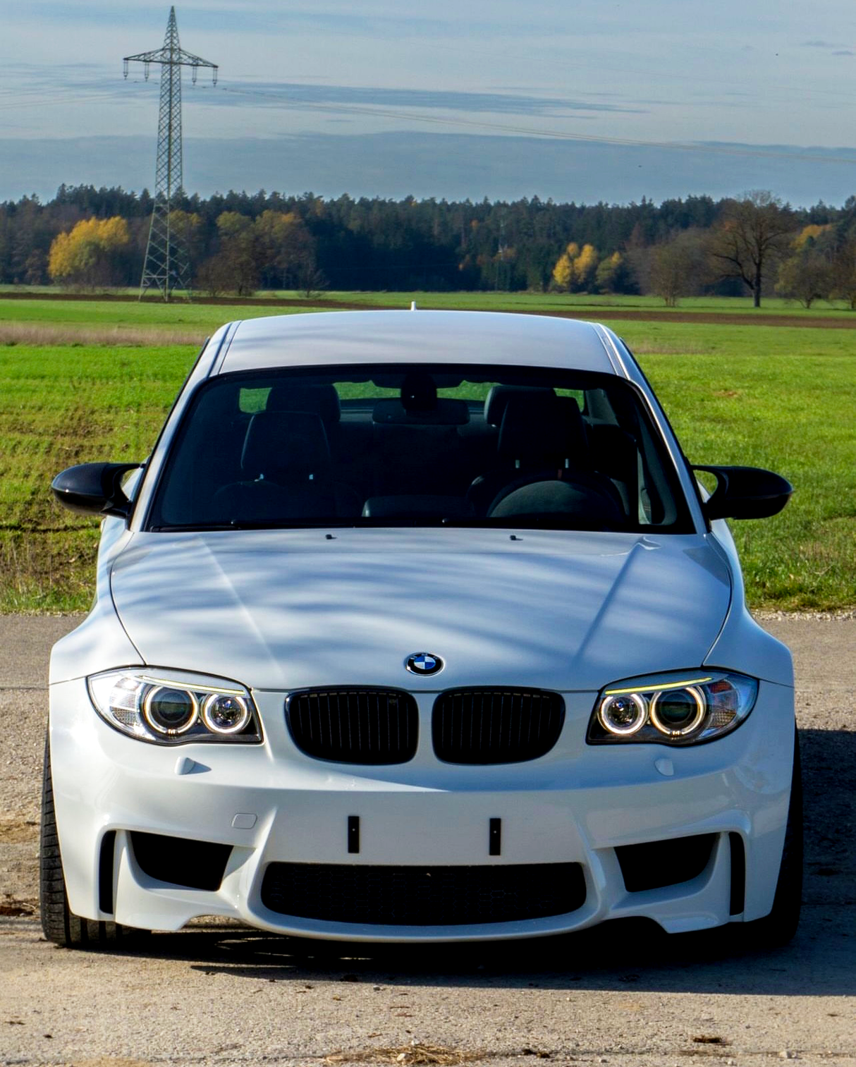 A white BMW car is parked on a rural road. It is facing forward with a power line in the background and surrounded by green fields and distant trees.