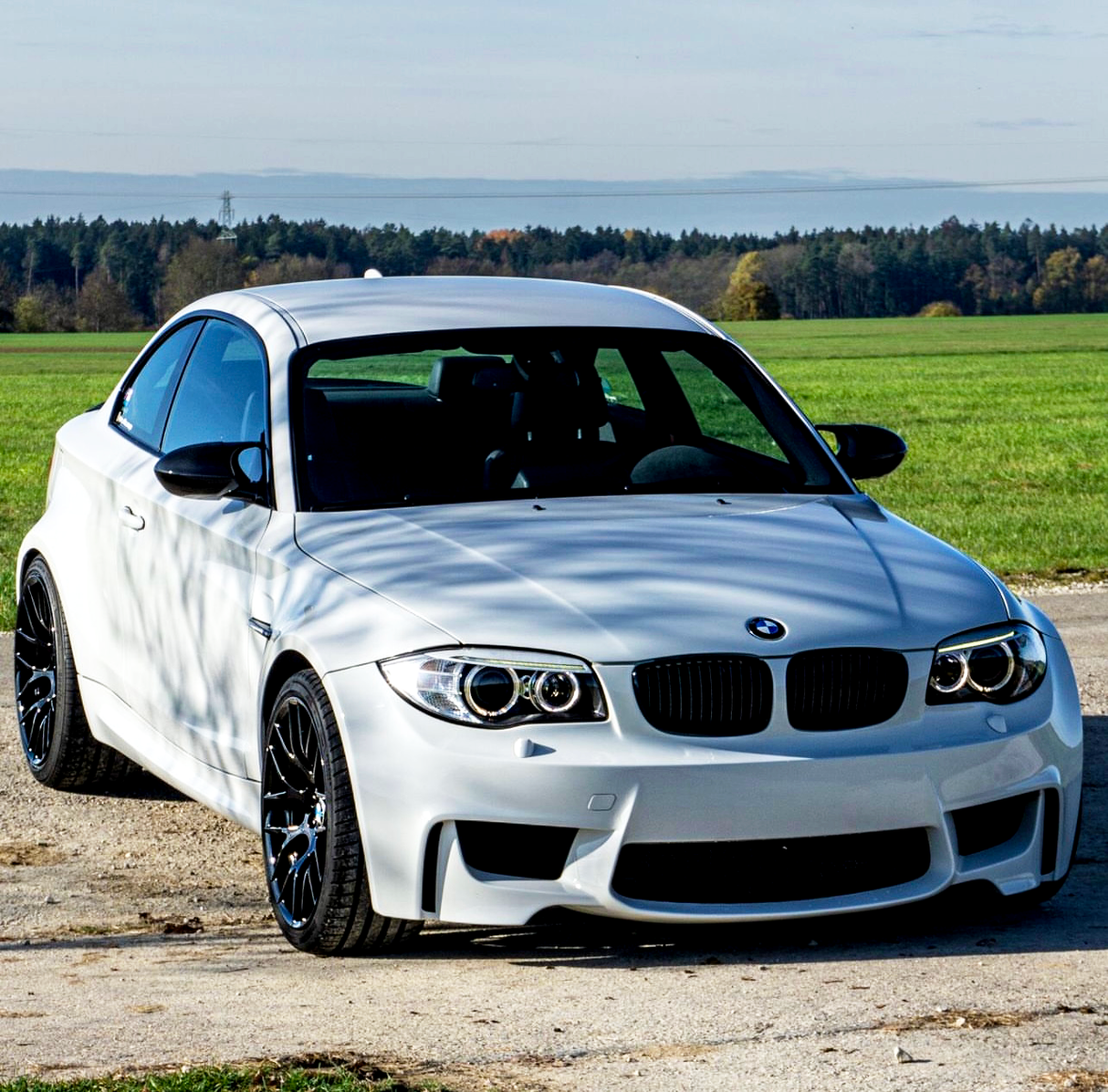 A white car is parked on a rural road, surrounded by green fields and a distant forest under a clear sky.