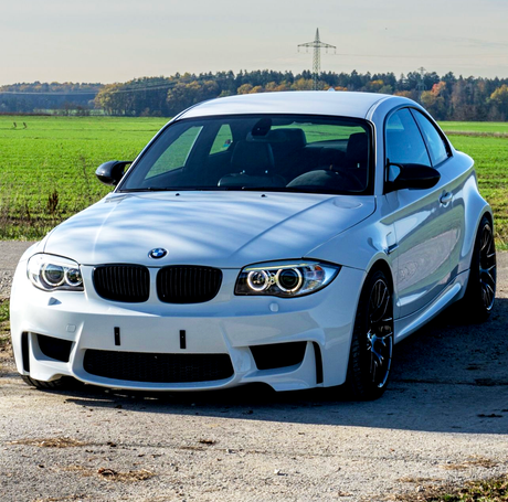 A white sports car is parked on a rural road with green fields and trees in the background under a clear sky.