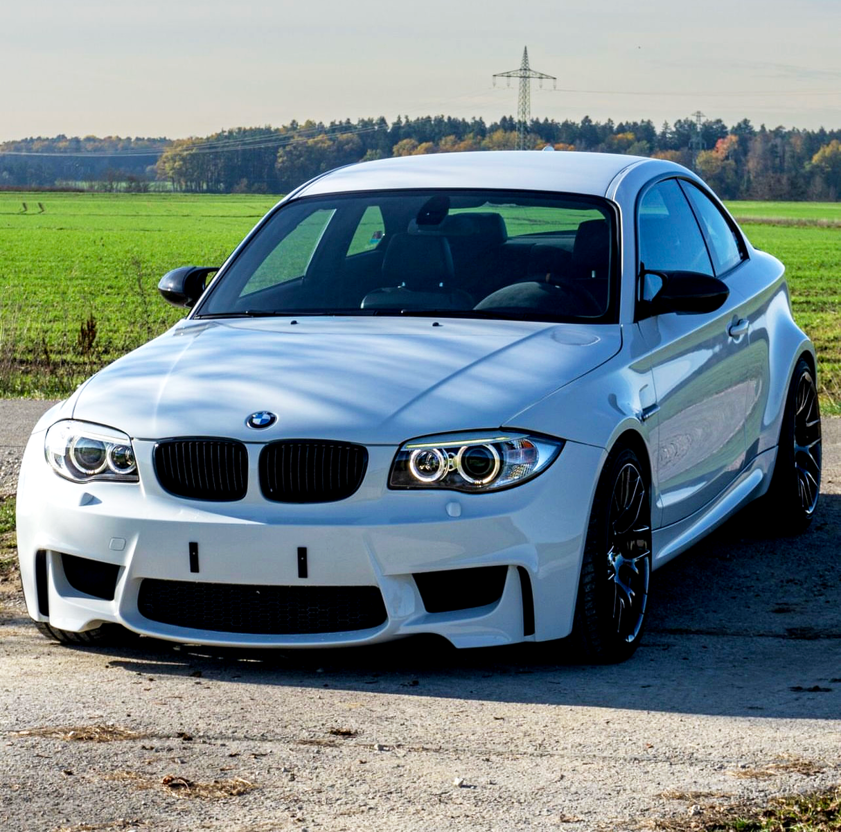 A white sports car is parked on a rural road with green fields and trees in the background under a clear sky.