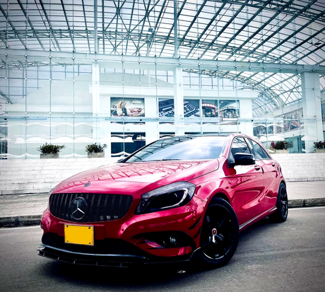 A red car with a distinctive emblem is parked on a street. It appears sleek and polished, in front of a modern glass building with a translucent roof structure.