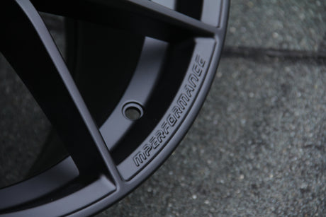 A black car wheel rim with the word "M PERFORMANCE" embossed on it, set against a textured grey surface.