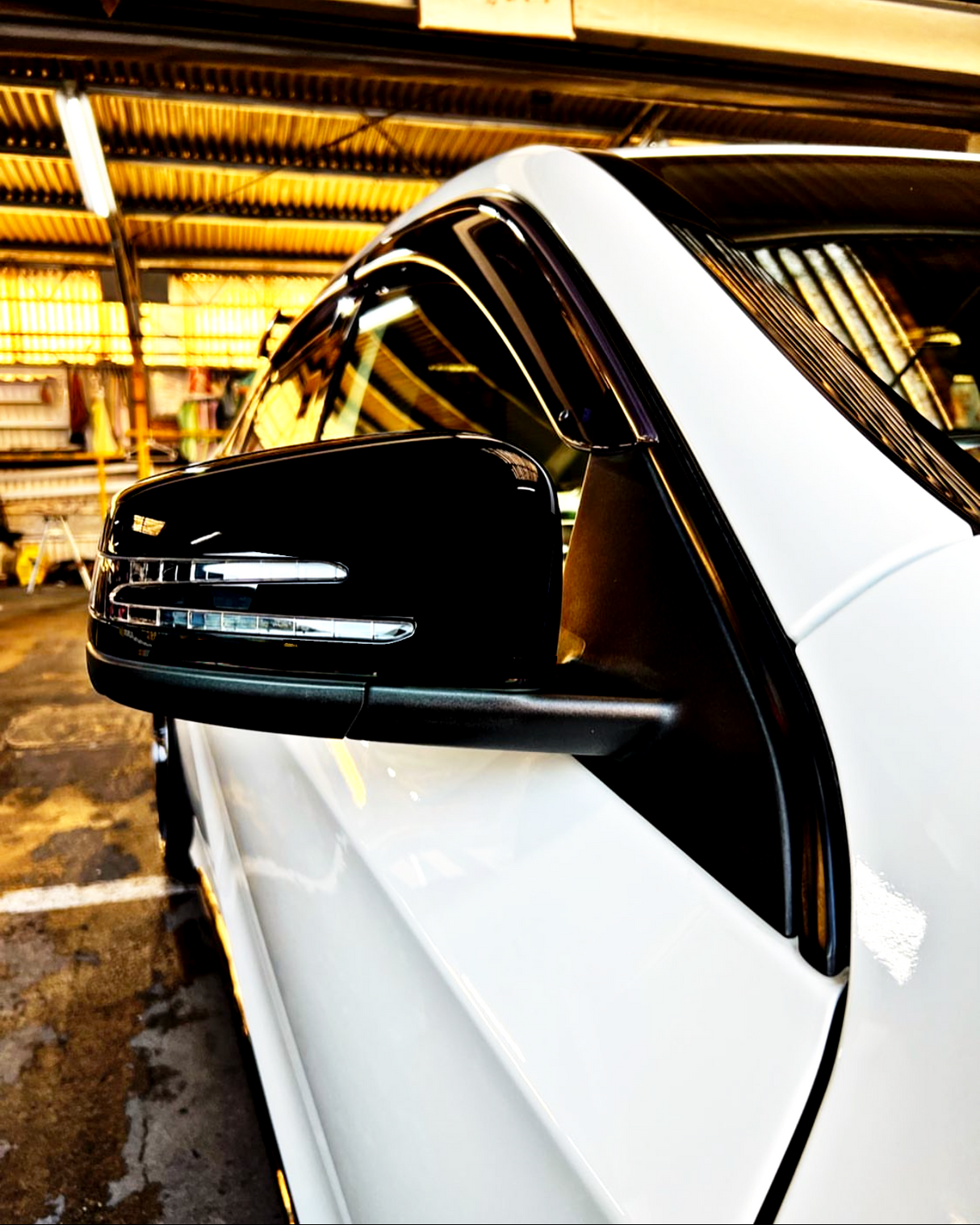 A sleek black side mirror reflects light, attached to a white vehicle, parked in a dimly lit garage with a corrugated metal roof.