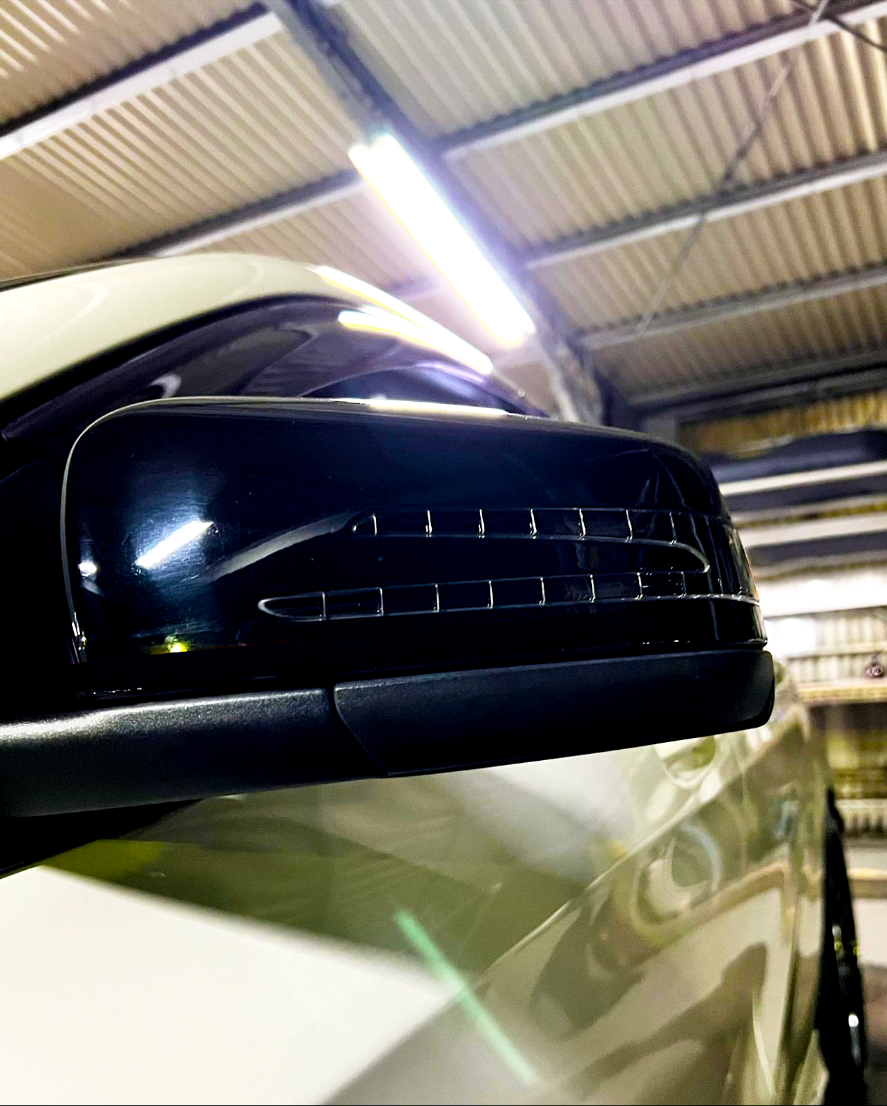 A car side mirror with integrated indicator lights is illuminated within a garage, where the ceiling features exposed metallic beams and fluorescent lighting.