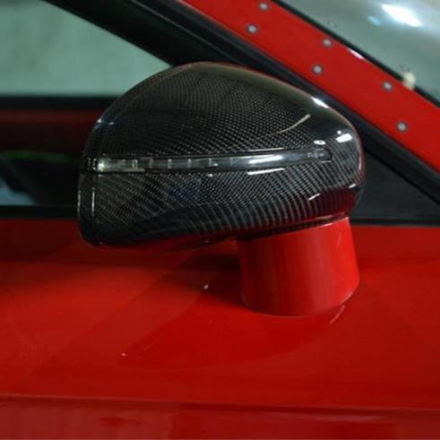 A carbon fiber car side mirror, featuring a sleek, aerodynamic design, is mounted on a vibrant red vehicle. The background is blurred, emphasizing the mirror's reflective surface.