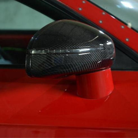 A carbon fiber car side mirror, mounted on a bright red vehicle, reflects light with a sleek, aerodynamic shape in an indoor setting.