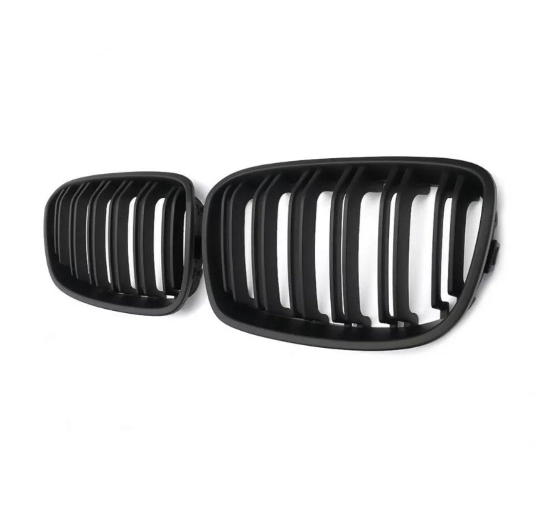 Black car grilles with vertical slats are positioned side by side, against a plain white background, emphasizing their design and structure.