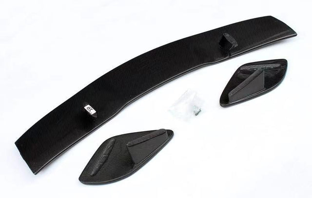 Carbon fiber car spoiler components lie on a white surface, consisting of a long curved piece and two smaller side elements. A small bag containing screws is nearby.