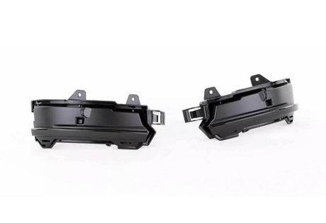 Two black plastic automotive parts, likely fog light housings, are positioned side by side facing outward. They feature mounting brackets and an angular design, set against a plain white background.