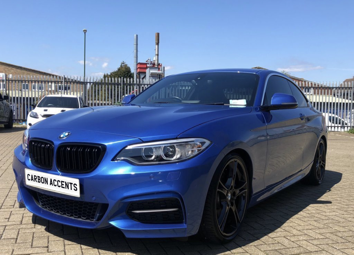 A blue sports car parked on a paved area, featuring a license plate reading "CARBON ACCENTS," surrounded by industrial buildings and fencing under a clear sky.
