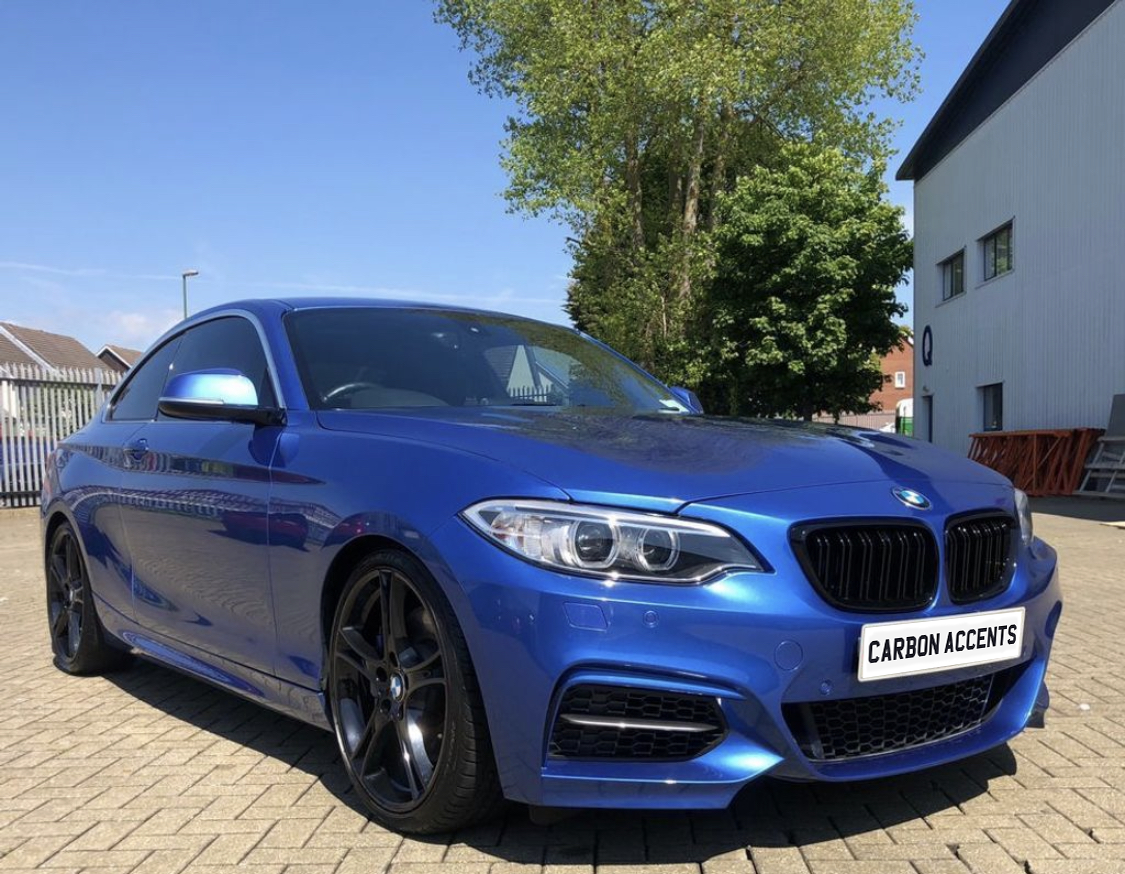 A blue car is parked in a sunny lot, surrounded by trees and buildings. The front license plate reads "CARBON ACCENTS."