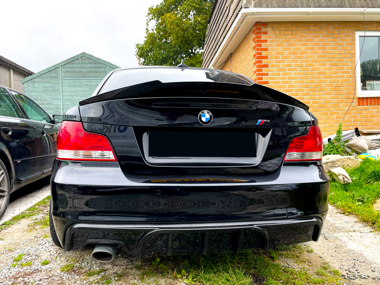 A black BMW car is parked on a gravel driveway beside a brick house and a green wooden shed, with its rear view showing distinct red taillights.