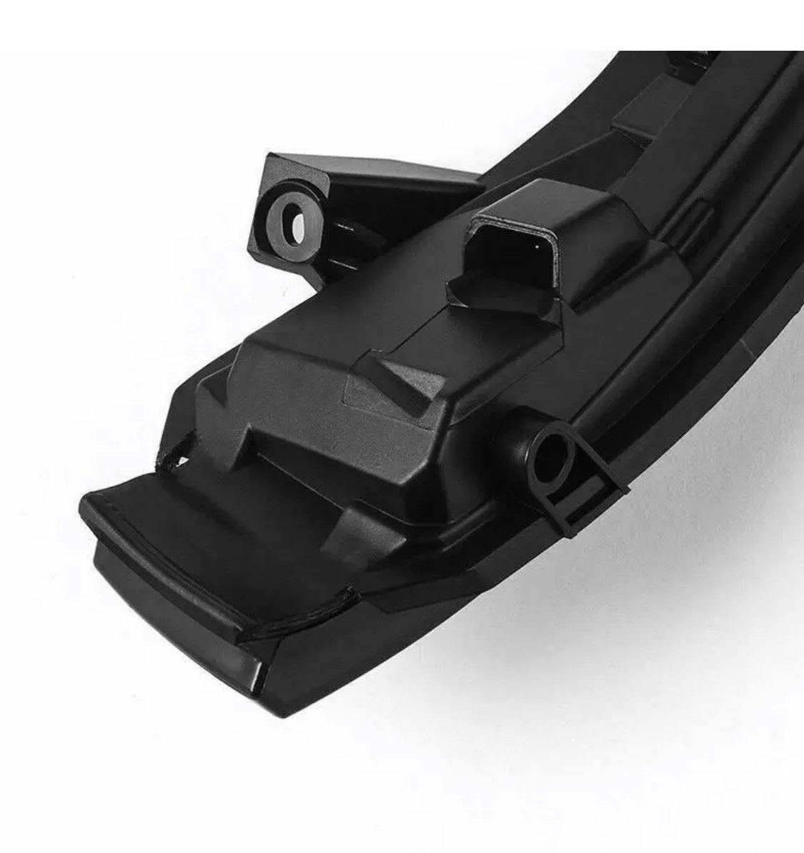 A black, angular, plastic automotive component with a rectangular connector port sits on a smooth, white background, featuring mounting points and a sleek, modern design.