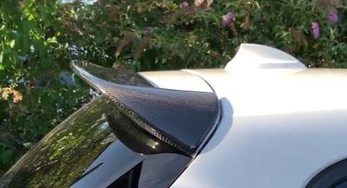 A carbon fiber car spoiler is mounted on a white vehicle under sunlight, with a background of green foliage and pink flowers.
