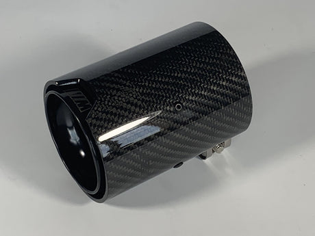 A cylindrical carbon fiber exhaust tip displays reflective surface details, resting on a solid white background. It features a small logo with stylized letters "MW" on the side.