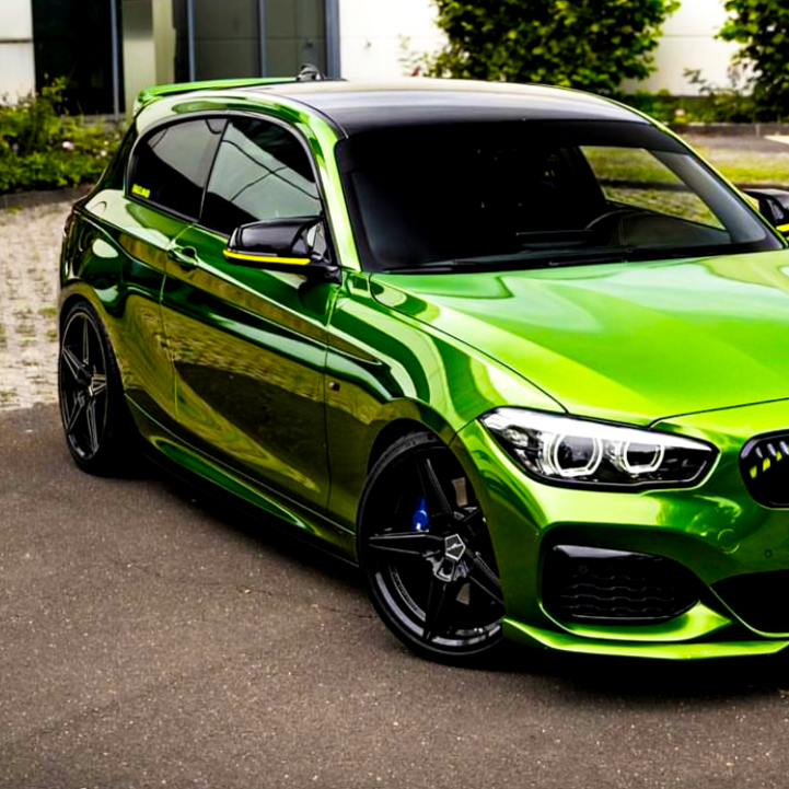 Green car parked on asphalt, featuring sleek black accents and tinted windows, with a building and greenery in the background.