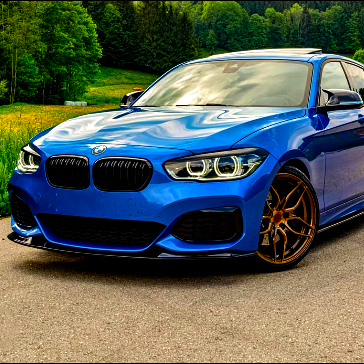 A blue car with gold wheels is parked on a road, surrounded by lush green trees and grass, under a cloudy sky.