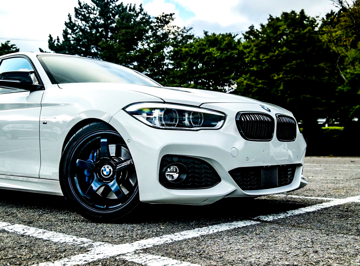 A white BMW car is parked on a paved area, featuring sleek, black alloy wheels and prominent headlights. Surrounding trees and a cloudy sky provide a natural backdrop.