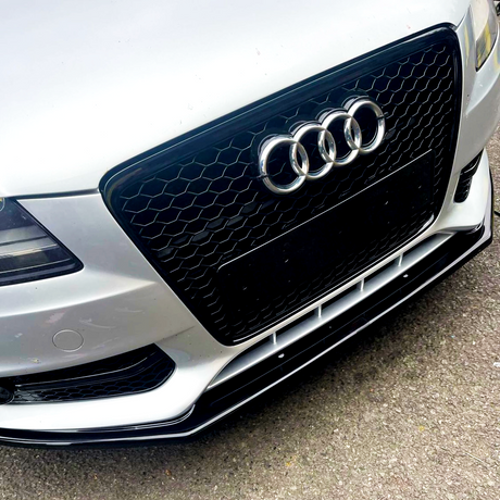 A silver car's front grill with a honeycomb pattern displays the Audi logo, consisting of four interlocking rings, against a concrete background.