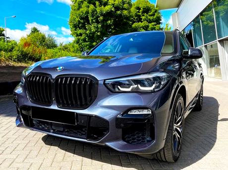 A sleek, dark SUV with prominent grille and illuminated headlights is parked on a paved driveway, near a modern building, surrounded by green trees under a sunny, blue sky.