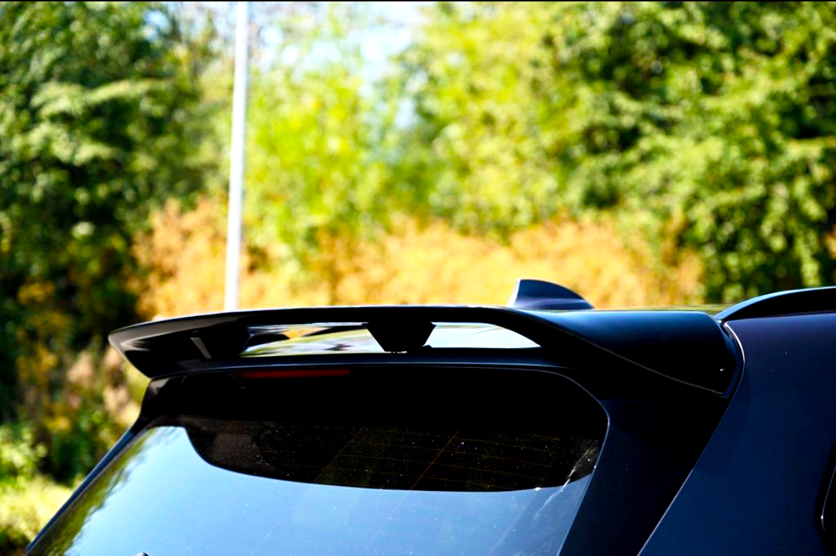 A black car spoiler is mounted on the rear of a vehicle, reflecting sunlight. The setting is outdoors, with blurred green foliage in the background.