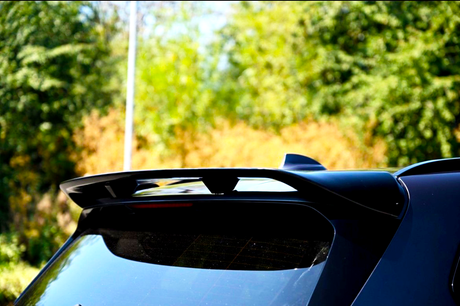 A black car spoiler is mounted on the rear of a vehicle, reflecting sunlight. The setting is outdoors, with blurred green foliage in the background.
