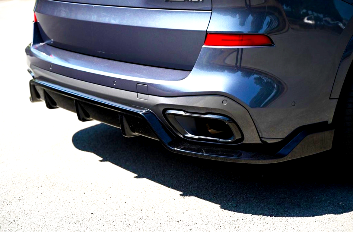 A dark metallic car's rear end, featuring dual exhaust tips and a diffuser, is parked on a sunlit asphalt surface. The vehicle's body reflects nearby buildings and trees.