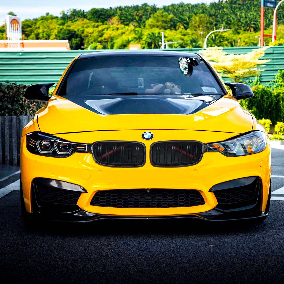 Yellow BMW car is parked, featuring a black hood, in a parking area with greenery and buildings in the background. A plush toy is visible inside on the dashboard.