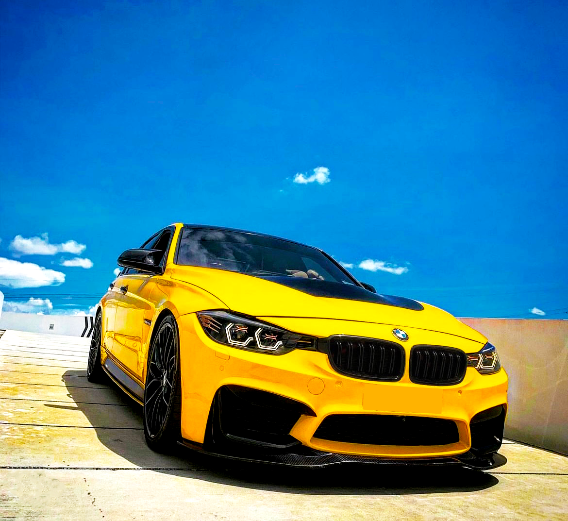 A yellow sports car is parked on a sunlit concrete surface. The sky is bright blue, with a few scattered clouds in the background.