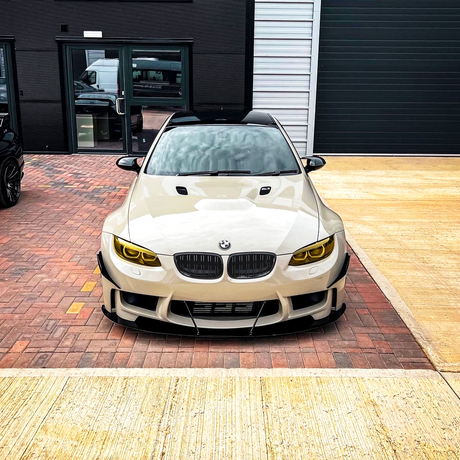 A white BMW car sits parked on a brick driveway in front of a modern building, featuring yellow-tinted headlights and a sleek, aggressive front design.