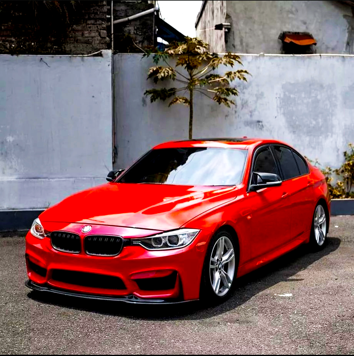 A shiny red sports car is parked on a paved area. It is positioned against a plain, gray concrete wall, with a small tree nearby and an adjacent building.