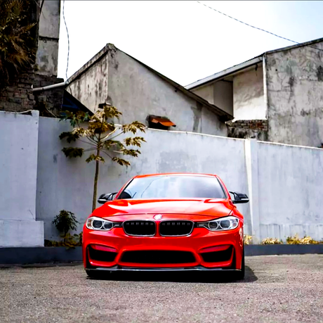 A red sports car is parked on the pavement, facing forward. It is situated in front of a gray wall with small plants, adjacent to weathered buildings.