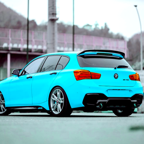 Blue car parked outdoors, showcasing its rear on gray pavement. Red taillights contrast with the bright color. In the background, a fenced area and blurred hilly terrain are visible.