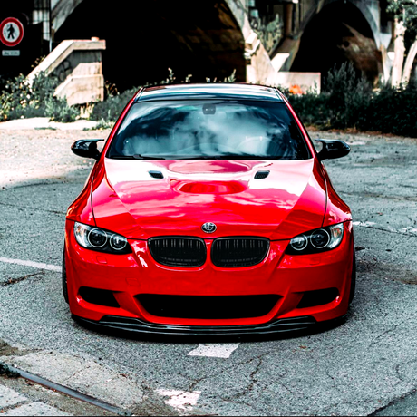 A red sports car is parked on a textured, paved area near a stone stairway and an underpass, with foliage in the background. The setting is urban and shaded.