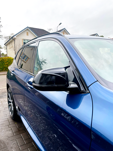 A blue car is parked on a wet driveway, showing a side mirror and window. It reflects nearby houses and a cloudy sky in a suburban neighborhood.