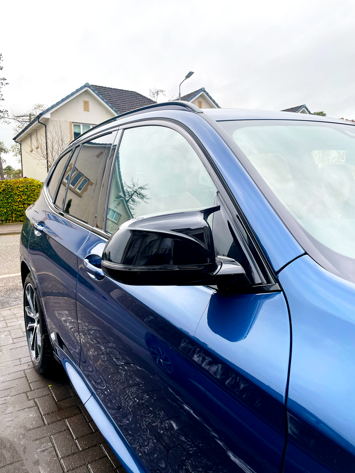 A shiny blue car is parked, reflecting nearby houses and trees on its surface, situated in a residential street with overcast skies.