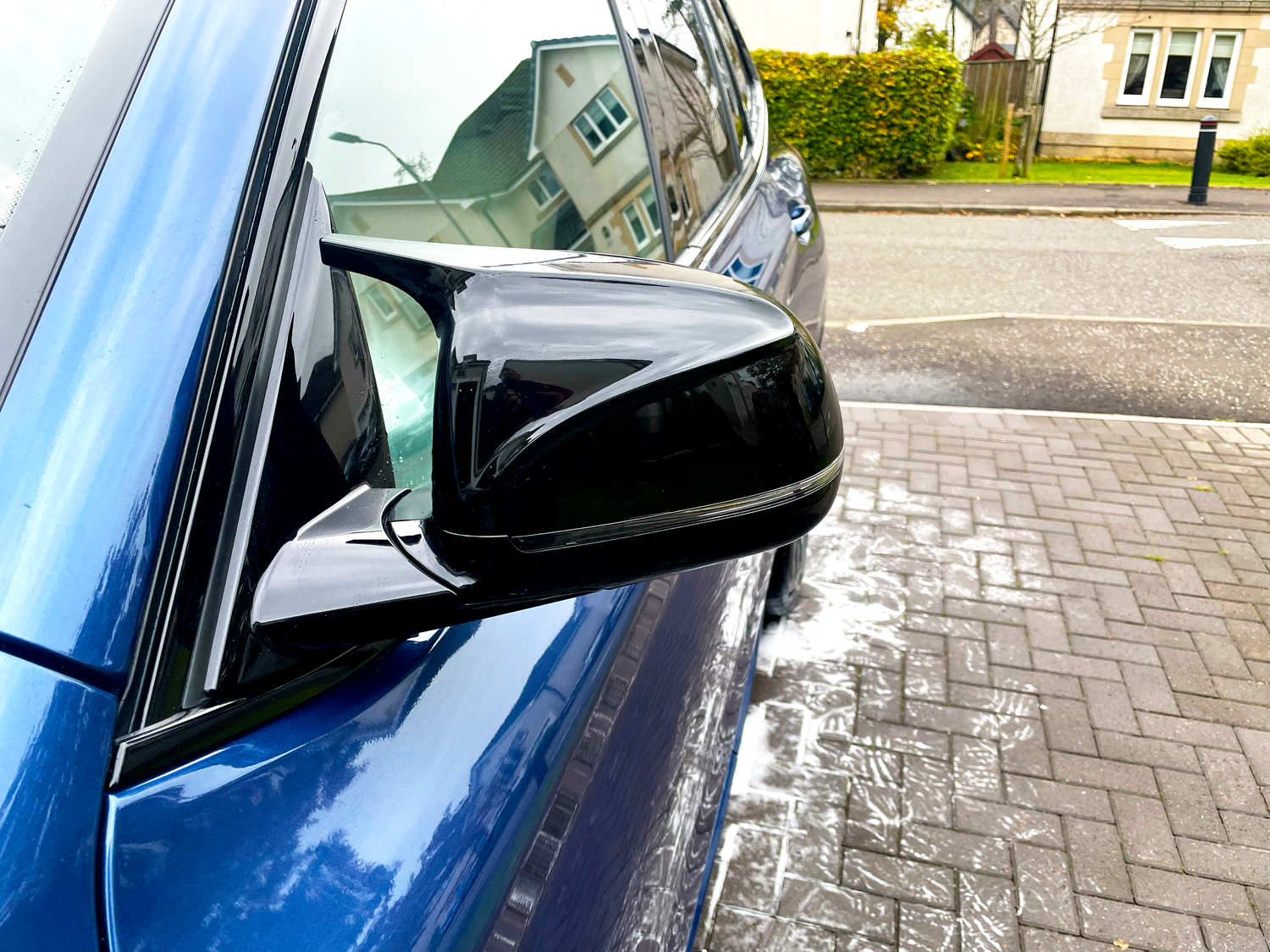 A shiny car side mirror reflects a suburban street, with soapy water on the brick driveway indicating a recent wash. Nearby, tidy houses and greenery create a tranquil setting.
