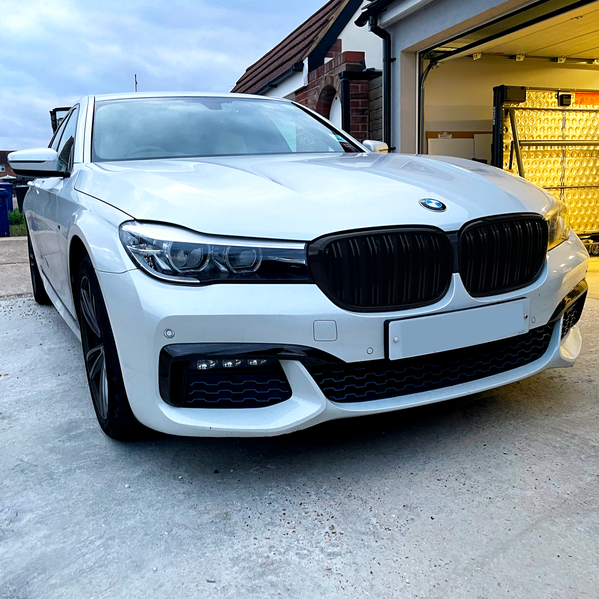 A white BMW sedan is parked in a driveway near an open garage with illuminated interiors, under a cloudy sky. The license plate is visible but blank.