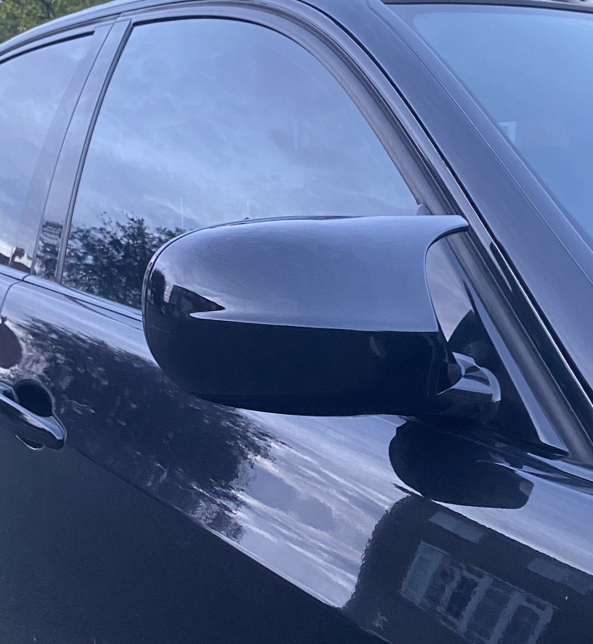 A car's side mirror reflects the surroundings, attached to a sleek, dark vehicle door. The background shows trees and sky, reflecting on the tinted windows and shiny surface.