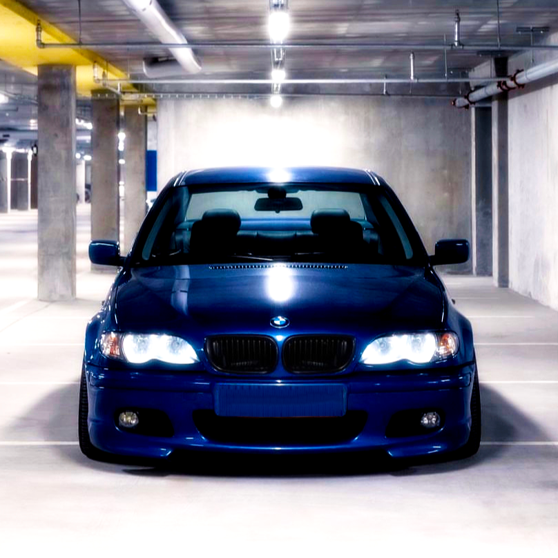 A blue car with headlights on is parked in an empty, dimly lit underground parking garage, surrounded by concrete pillars and overhead pipes.