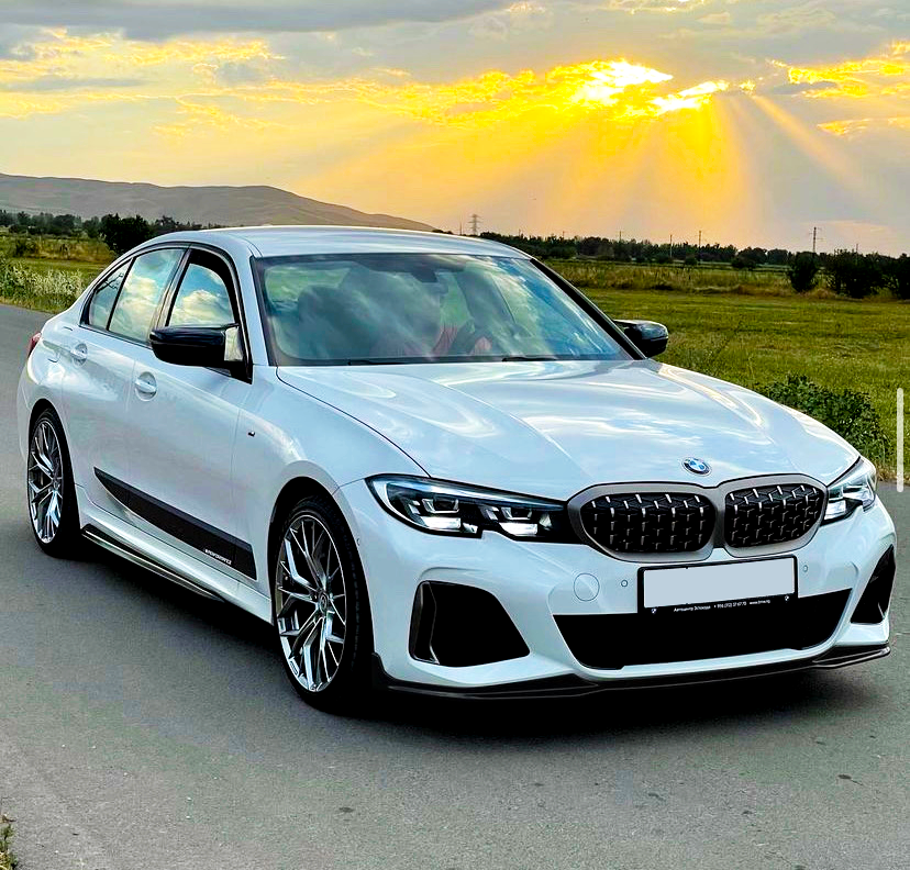 A white BMW sedan is parked on a road, with sunlight streaming through clouds in a grassy landscape. The license plate reads, "Premium Selection - 11011210 g73/11476 Omringes."