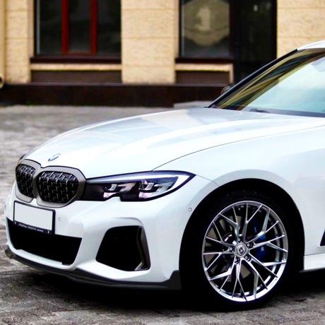 A white BMW car is parked on a cobblestone surface in front of a beige building with windows. The focus is on the front left side of the vehicle.